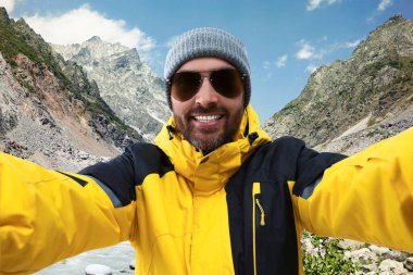 Şapkalı ve güneş gözlüklü gülen adam dağlarda selfie çekiyor.