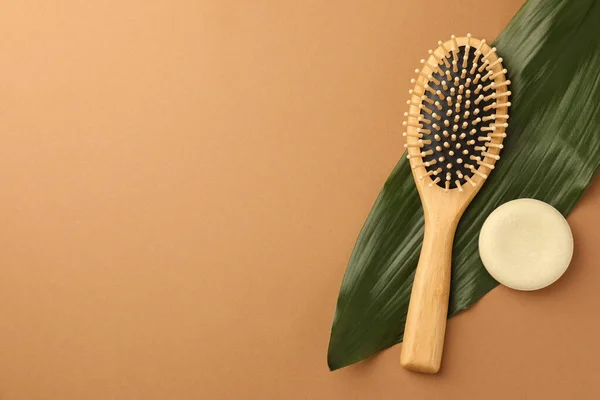 Wooden hairbrush, solid shampoo and green leaf on light brown background, flat lay. Space for text