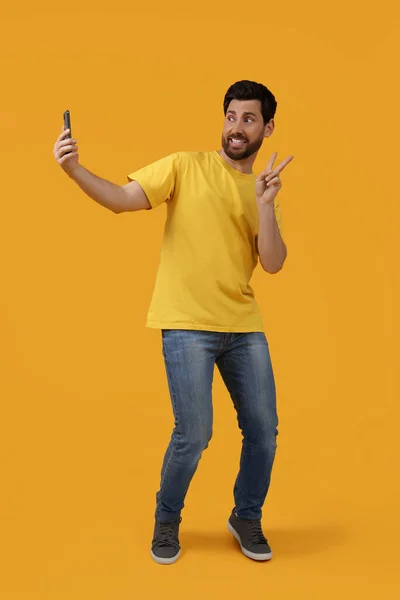 微笑的男人拿着智能手机自私自利 在黄色的背景上展示和平的标志 — 图库照片