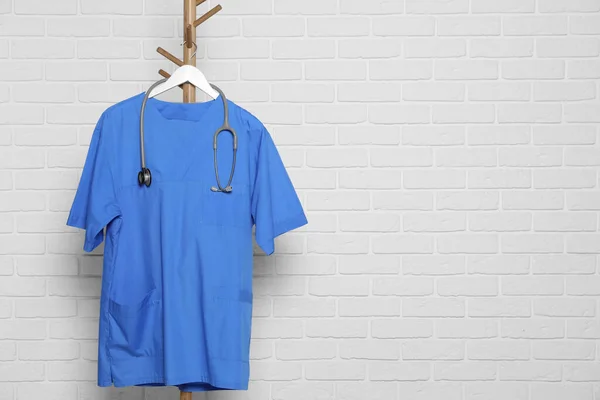 Blue medical uniform and stethoscope hanging on rack near white brick wall. Space for text