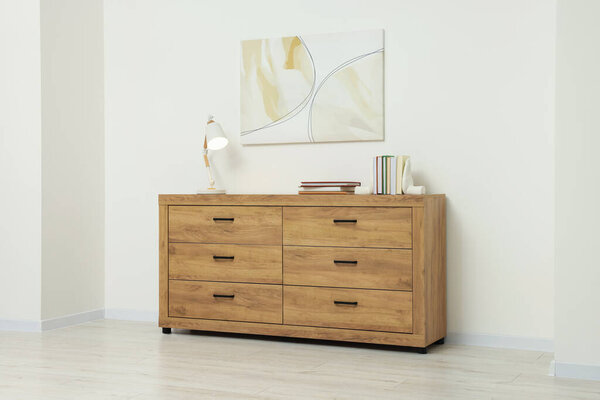 Wooden chest of drawers, lamp and beautiful picture on white wall indoors. Interior design