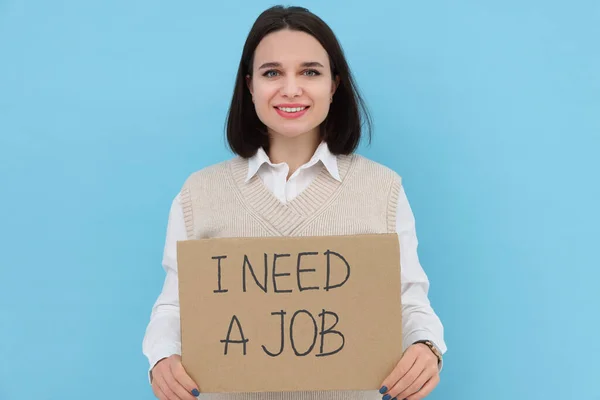 快乐的年轻失业妇女举着标语 上面写着 我需要一份浅蓝色背景的工作 — 图库照片