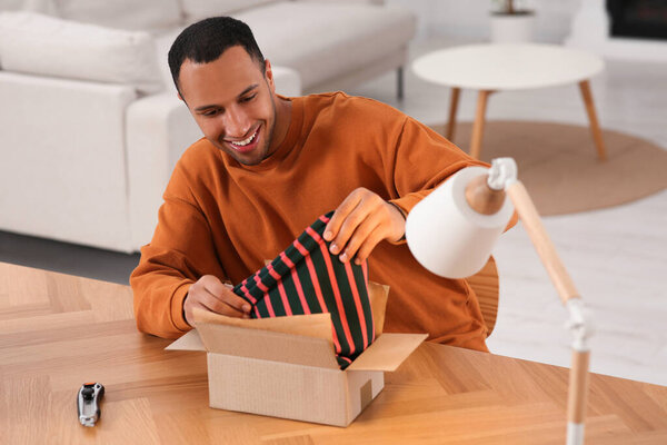 Happy young man opening parcel at table indoors. Internet shopping