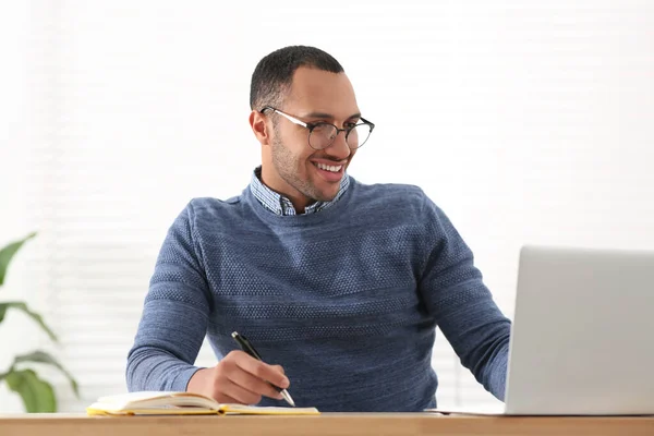 木製のテーブルでラップトップで作業している間にノートパソコンで書くアフリカ系アメリカ人の男性を笑顔 — ストック写真
