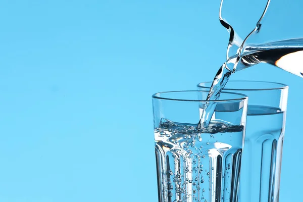 Pouring water from jug into glass on light blue background, closeup. Space for text