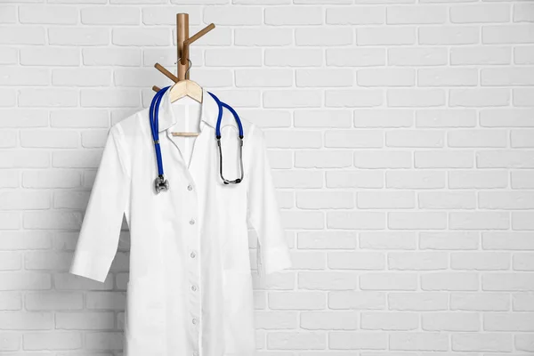 Medical uniform and stethoscope hanging on rack near white brick wall. Space for text