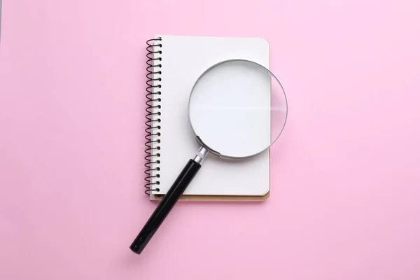 Magnifying glass and notebook on pink background, top view