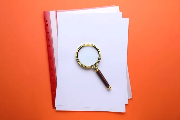 Magnifying glass and folder with paper sheets on orange background, top view