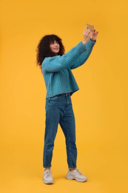 Güzel genç bir kadın turuncu arka planda akıllı telefonuyla selfie çekiyor.