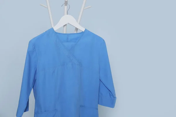 Blue medical uniform hanging on rack against light grey background. Space for text