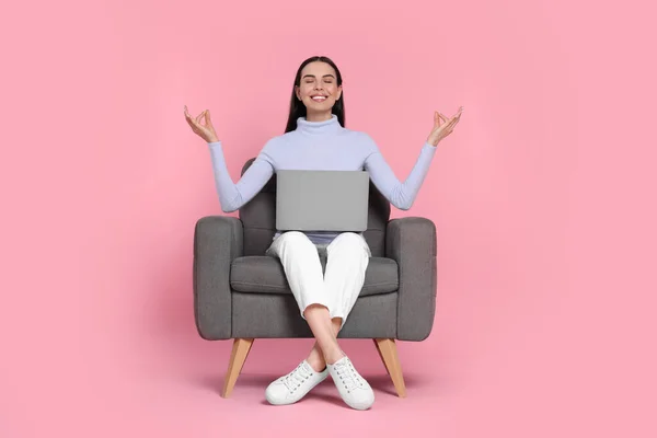 Happy woman with laptop sitting in armchair and meditating on pink background