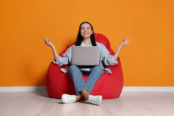 Happy woman with laptop sitting on beanbag chair and meditating near orange wall