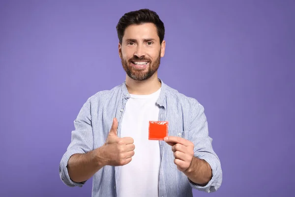 紫色の背景に親指を示すコンドームを持つ幸せな男 安全なセックス — ストック写真