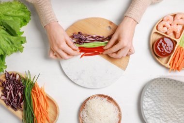Lezzetli börek yapıyorum. Beyaz ahşap masada taze sebzeleri pirinç kağıdına saran kadın.