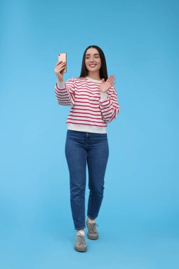 Gülümseyen genç bir kadın açık mavi arka planda akıllı telefonuyla selfie çekiyor.