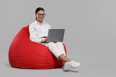 Dizüstü bilgisayarlı mutlu kadın açık gri arka planda puf koltukta oturuyor.