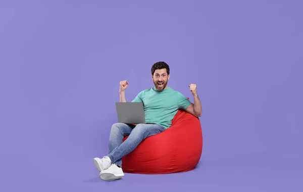 情绪激动的男人 带着笔记本电脑坐在紫色背景的豆袋椅上 — 图库照片