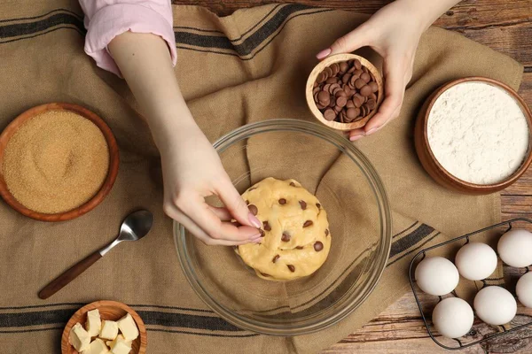 Cooking sweet cookies. Woman adding chocolate chips to dough at wooden table, top view