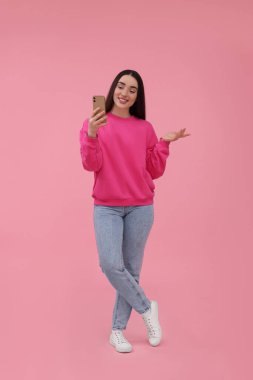 Gülümseyen genç kadın pembe arka planda akıllı telefonuyla selfie çekiyor.