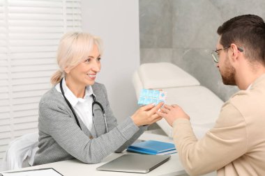 Doktorun elinde ilaç kutusu ve klinikteki beyaz masada danışmanlık yapan hasta var.