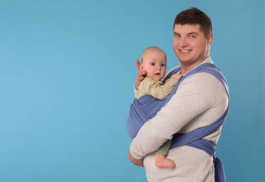 Father holding his child in sling (baby carrier) on light blue background. Space for text clipart