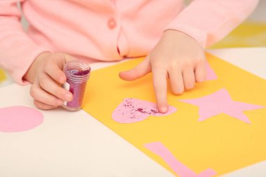 Girl using colorful glitter while making paper card at desk in room, closeup. Home workplace