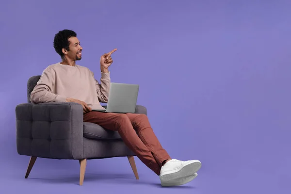 ノートパソコンを持つ幸せな男アームチェアに座って紫色の背景に何かを指している テキストのスペース — ストック写真