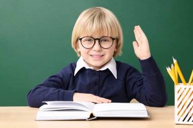 Küçük mutlu okul çocuğu masa başında karatahtanın yanında kitaplarla el kaldırıyor.