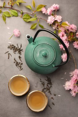 Traditional ceremony. Cups of brewed tea, teapot and sakura flowers on grey table, flat lay clipart