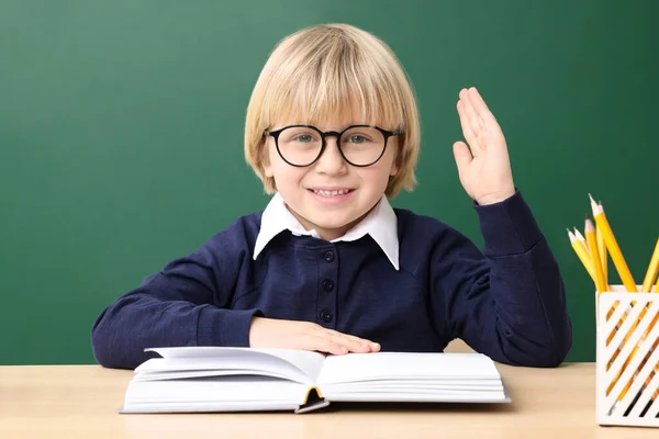 幸せな小さな学校の子供が手を上げながら 机の近くに本と一緒に座って黒板 — ストック写真