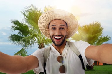Hasır şapkalı gülümseyen genç adam otelde selfie çekiyor.