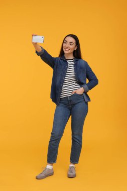 Gülümseyen genç kadın sarı arka planda akıllı telefonuyla selfie çekiyor.