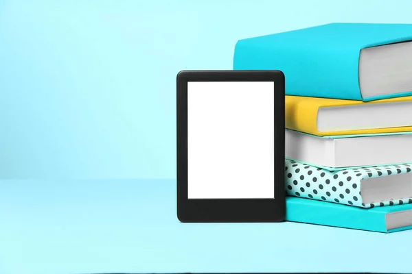 Modern e-book reader and stack of hard cover books on light blue background. Space for text
