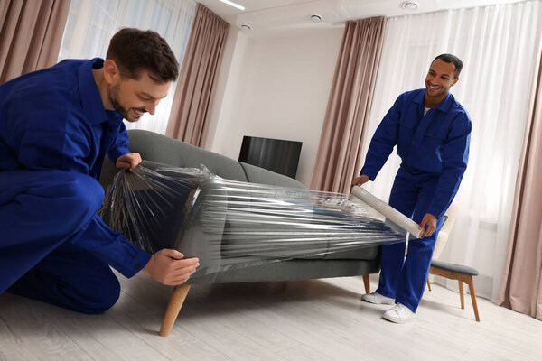 Male movers with stretch film wrapping sofa in new house