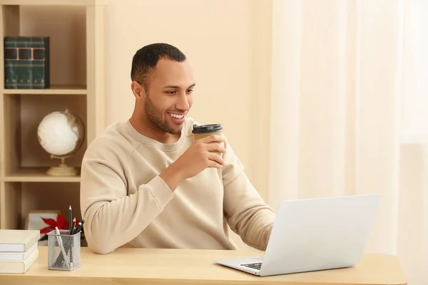 在房间内的木制桌旁 用咖啡在笔记本电脑旁度过时光的非洲裔美国人微笑 — 图库照片