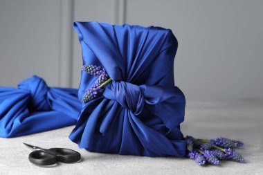 Furoshiki technique. Gifts packed in blue silk fabric, muscari flowers, ribbon and scissors on light grey table clipart