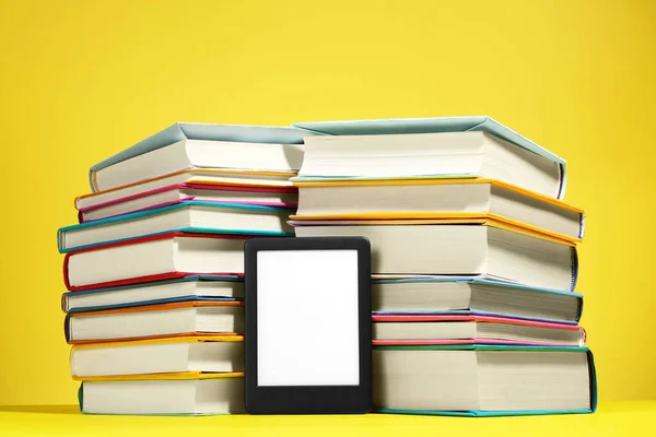 Modern e-book reader and stacks of hard cover books on yellow background