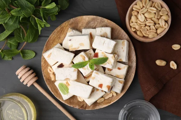 stock image Pieces of delicious nutty nougat, peanuts, mint and honey on gray wooden table, flat lay