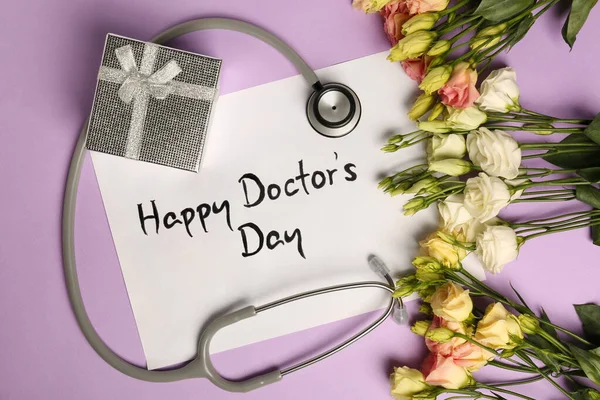 Card with phrase Happy Doctor\'s Day, stethoscope, gift box and flowers on purple background, flat lay