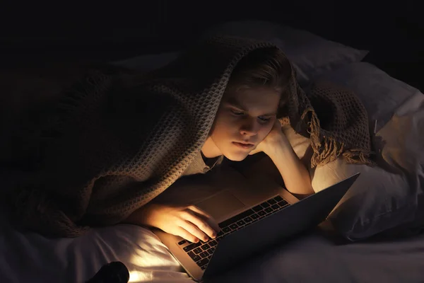 Teenage boy using laptop under blanket on bed at night. Internet addiction