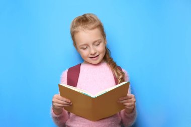 Sırt çantalı mutlu küçük kız açık mavi arka planda kitap okuyor.