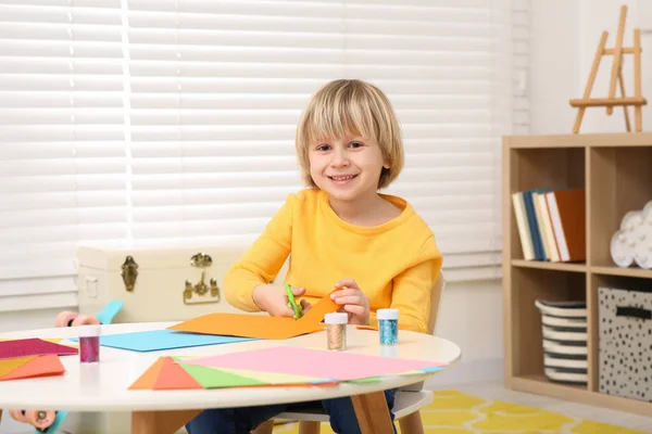 かわいい男の子が部屋の机でカラフルな紙を切っている ホームワーク — ストック写真