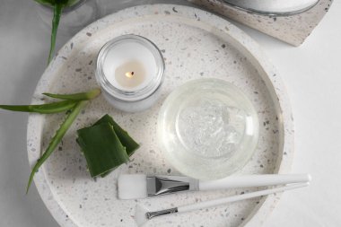 Kavanoz dolusu doğal jel, yanan mum, makyaj fırçaları ve beyaz masada taze aloe yaprakları.