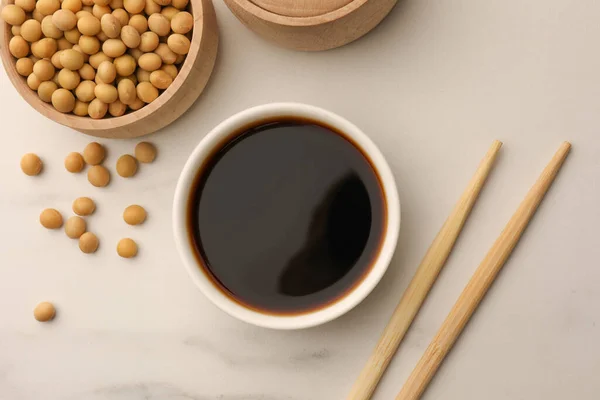 Stock image Soy sauce in bowl, soybeans and chopsticks on white marble table, flat lay