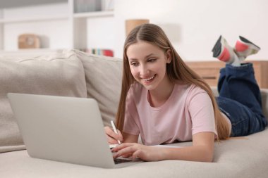 Çevrimiçi öğrenme. Genç kız evdeki kanepede dizüstü bilgisayarın yanında özet yazıyor.