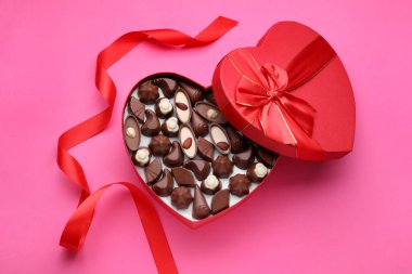 Heart shaped box with delicious chocolate candies and ribbon on pink background, flat lay