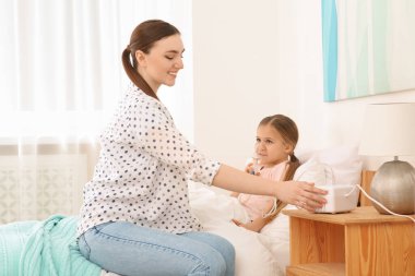 Anne hasta kızına yatak odasında nebulizör solumasında yardım ediyor.