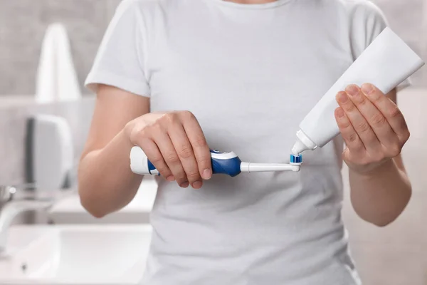 Woman squeezing toothpaste from tube onto electric toothbrush in bathroom, closeup