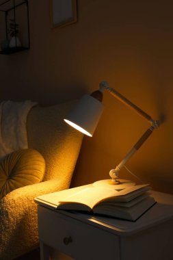 Modern masa lambası ve karanlık odadaki koltuğun yanındaki beyaz dolapta açık bir kitap.
