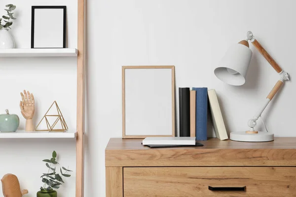 Stylish modern desk lamp, books and frame on wooden chest of drawers near white wall indoors